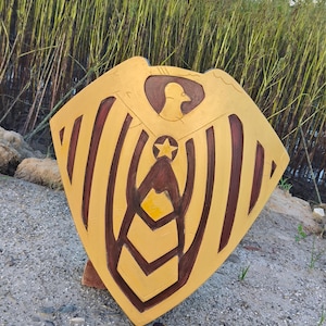 May include: A golden shield with brown accents, featuring a central star and bird emblem. The shield has a geometric design with vertical and angled lines. The shield is resting on a sandy surface with tall grass in the background.