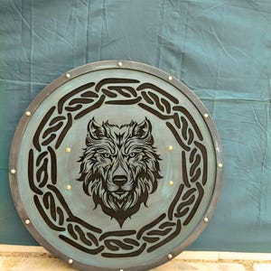 Könnte beinhalten: Ein runder Schild mit einem Wolfskopf-Design. Der Schild hat einen türkisfarbenen Hintergrund mit einem schwarzen Wolfskopf in der Mitte, umgeben von einem schwarzen keltischen Knotenmuster. Der Schild ist mit einem dunklen Metallband und kleinen Nieten versehen.