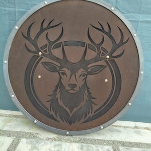 Könnte beinhalten: Ein runder, brauner Schild mit einem silberfarbenen Metallrand. Der Schild zeigt eine detaillierte schwarze Illustration eines Hirschkopfes in einem kreisförmigen Design. Nieten sichern den Rand. Ein dekorativer Gegenstand.