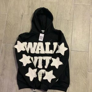 Puede incluir: Sudadera con capucha negra con texto blanco y detalles de estrellas. La palabra "WALK" está en el pecho y "VIT" debajo. Estrellas blancas cosidas en las mangas. Una etiqueta cuelga de la capucha.