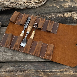 May include: A brown leather tool roll with several wood carving tools. The roll is open, displaying the tools in their individual slots. The tools have wooden handles and metal blades. The roll is tied with a leather cord.