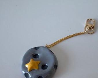 May include: A handmade keychain featuring a gray, moon-shaped pendant with black craters and a gold star. The pendant is attached to a gold chain and clasp, creating a celestial-themed accessory.