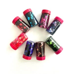 May include: A collection of small, clear plastic jars with red lids, each filled with colorful flower-shaped glitter. The glitter colors include red, pink, blue, purple, and green, creating a vibrant display. The jars are arranged in a circular pattern.