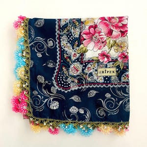 May include: A square, navy blue scarf with a floral pattern and a colorful, fringed edge. The scarf features a large pink flower design on a white background, with the word "ERIPEK" printed on a small label.