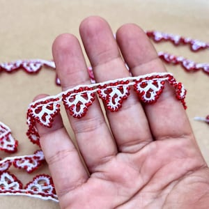 May include: A length of white crocheted lace trim featuring a repeating triangular pattern edged with small, vibrant red beads. The delicate textile design consists of interconnected loops and knots, creating a decorative border with a consistent motif.