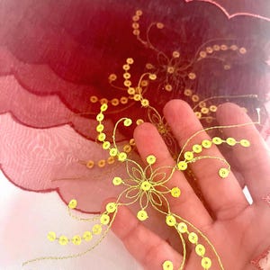 May include: Close-up of a sheer, burgundy fabric with a scalloped edge. Gold-colored thread and sequins are sewn onto the fabric in a floral and vine-like pattern. The fabric is held in a hand.
