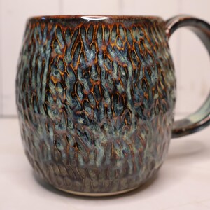 May include: A ceramic mug with a textured, organic surface featuring a reactive glaze in shades of deep blue, teal, and copper brown. The mug has a rounded, barrel-like body and a large, curved handle that matches the exterior color palette.