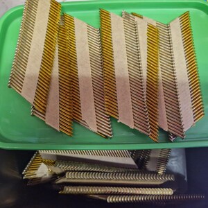 May include: A green plastic tray contains angled strips of collated nails, with a gold and silver appearance. The nails are held together with tan paper tape. More nails are visible below the tray.