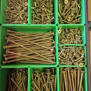 May include: An assortment of gold-coloured screws, varying in size and shape, stored in green plastic compartments. The screws are arranged in a grid, displaying different types suitable for various construction tasks.
