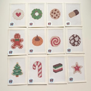 May include: A set of twelve needlepoint canvases featuring holiday-themed designs including a gingerbread person, a candy cane, a Christmas tree, cookies, and a star. Each rectangular canvas has a white border and a small circular SF logo.