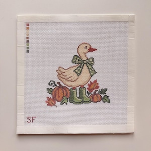 Peut inclure: Un motif de point de croix sur tissu blanc représentant un canard jaune clair portant un nœud vert à pois et des bottes de pluie vertes. Le canard est entouré de citrouilles orange et de feuilles d'automne. Les lettres SF sont brodées en bas.