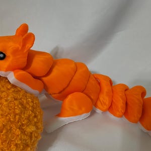 May include: An orange plush squirrel with a segmented body and white accents. The squirrel is perched on a textured, orange, round object. The squirrel has black eyes and a detailed head.