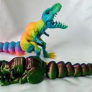 Könnte beinhalten: Zwei bewegliche Dinosaurier-Spielzeuge. Eines ist ein Regenbogen aus Farben, das andere ist grün und bronze. Der Regenbogen-Dinosaurier steht, während der grüne liegt. Beide haben offene Münder und detaillierte Merkmale.