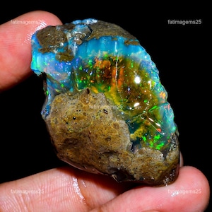 May include: A raw, unpolished opal gemstone featuring a mix of earthy brown rock matrix and vibrant, translucent sections displaying iridescent flashes of neon blue, bright green, and fiery orange colors against a dark, natural background.
