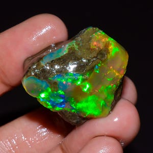 May include: A raw, uncut opal gemstone featuring vibrant flashes of neon green, blue, and orange play-of-color against a translucent, earthy matrix. The stone has a wet, polished appearance and includes the text fatimagems25.