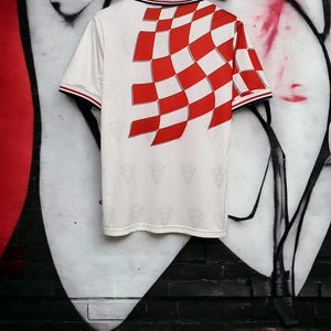 May include: White soccer jersey with a red and white checkered design on the back. The jersey has a polo collar with red, white, and blue trim. The sleeves have red and blue trim. The jersey is hanging on a wooden hanger.