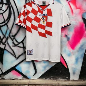 May include: White football shirt featuring a red and white chequered pattern across the shoulder and chest. It has a collar with blue and red trim, the Lotto logo, and the Croatian Football Federation emblem. The shirt is hanging on a wooden hanger.