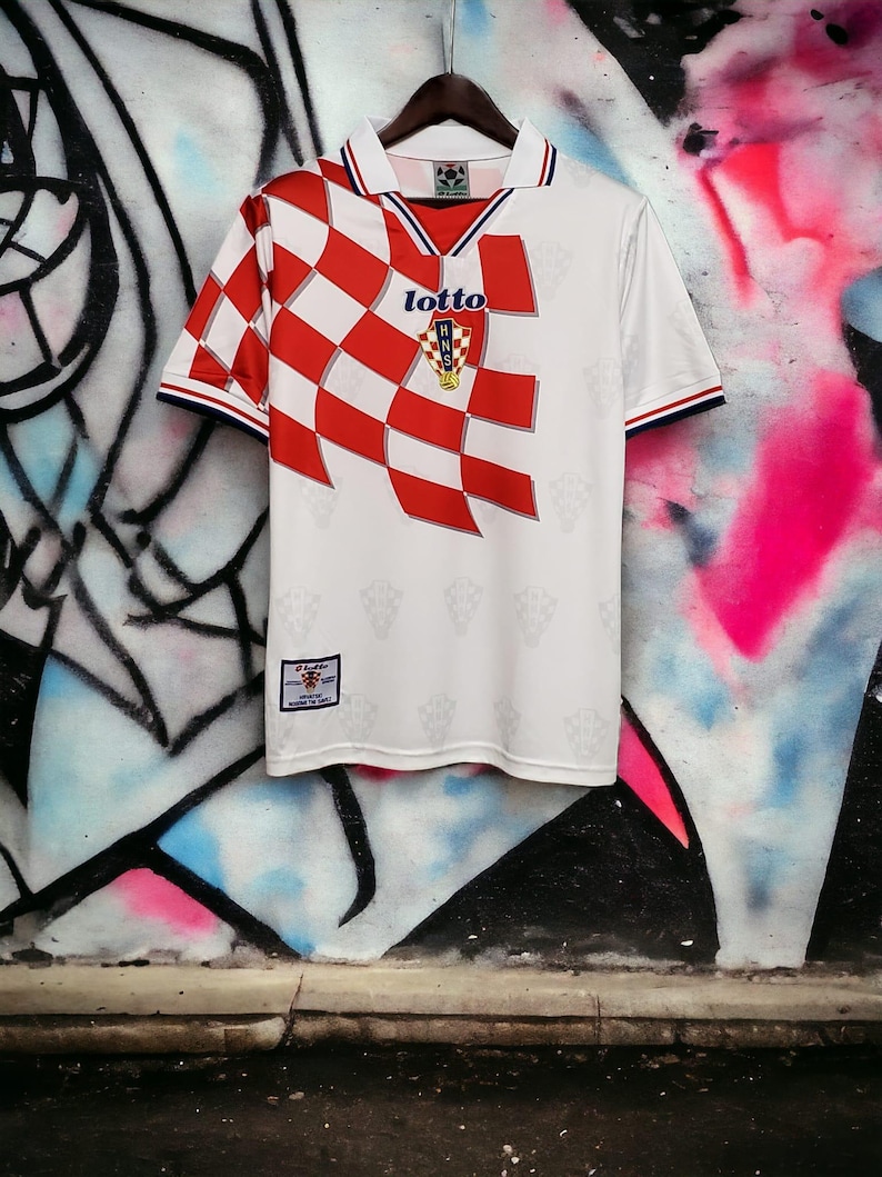 May include: White soccer jersey with a red and white checkered design on the left shoulder and chest. The jersey has a collar with blue and red trim, and the Lotto logo is visible. The Croatian Football Federation emblem is on the chest.