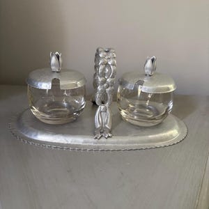 May include: A silver-toned serving set with two glass jars, each topped with a tulip-shaped lid. A matching floral-design handle and an oval tray complete the set. The jars are clear, and the set is on a gray surface.