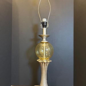 May include: A decorative table lamp with a gold-colored base and a spherical amber-colored glass shade. The lamp has a silver-colored metal frame for the lampshade and a gold-colored finial at the top. The lamp is plugged into a wall outlet.