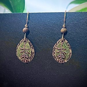 May include: A pair of handcrafted earrings featuring a leaf design. The earrings have a teardrop shape with a green and bronze color scheme. Each earring has a small, round bead at the top and a hook for the ear.