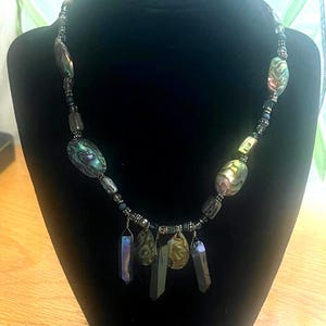 Può includere: Una collana di perline con perline iridescenti di conchiglia di abalone e ciondoli di cristallo grigio scuro. La collana presenta una varietà di forme e dimensioni di perline, infilate su un cordoncino scuro. La collana è esposta su un manichino nero.