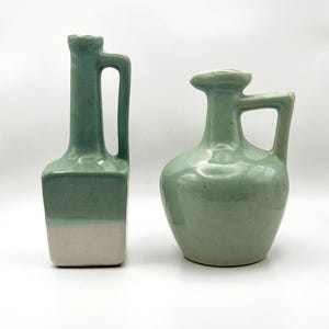 Może przedstawiać: Dwa ceramiczne wazony w miękkim odcieniu zieleni. Jeden jest prostokątny z uchwytem, ​​drugi jest zaokrąglony z uchwytem. Spód prostokątnego wazonu jest jasno-beżowy. Oba wazony mają błyszczące wykończenie.