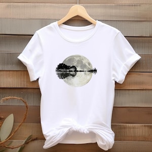 Könnte beinhalten: Weißes T-Shirt mit einem Vollmond-Motiv, einer Baumsilhouette und deren Spiegelung. Auf der rechten Seite der Mondspiegelung befindet sich eine Stadtsilhouette. Das T-Shirt hängt an einem Holzbügel.