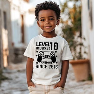 Könnte beinhalten: Ein Kind trägt ein weißes T-Shirt mit einem schwarzen Grafik-Controller und dem Text "LEVEL 10 UNLOCKED AWESOME SINCE 2016". Das Kind lächelt und steht draußen.