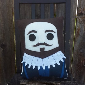 May include: A decorative pillow featuring a stylized face with a mustache, beard, and ruff collar. The pillow has a brown background with a white face, black eyes, and a blue and black outfit. The pillow is sitting on a black chair.