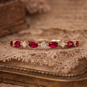 Op de afbeelding: Een gouden ring met afwisselend ovaal geslepen rode edelstenen en kleine, ronde, heldere edelstenen. De ring ligt op een delicate, kantachtige ondergrond met een warme, neutrale achtergrond.