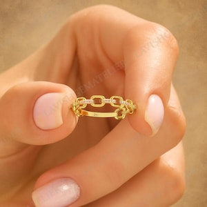 Peut inclure: Une bague en or avec des maillons de chaîne et de petites pierres transparentes serties dans les maillons. La bague est tenue entre deux doigts et un pouce. C'est un bijou délicat.