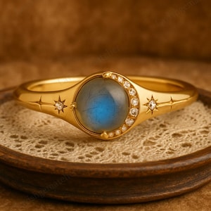 Puede incluir: Anillo dorado con una piedra preciosa azul redonda en el centro, rodeada de pequeños diamantes y motivos de estrellas. El anillo está sobre una superficie texturizada. El diseño es elegante y de temática celestial.
