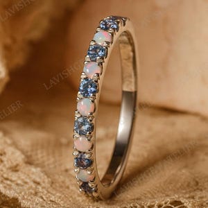 Puede incluir: Un anillo de plata con una fila de piedras preciosas azules y blancas alternas. Las piedras preciosas están engastadas en un engaste de canal y el anillo tiene un acabado pulido. El anillo se muestra sobre una superficie texturizada.