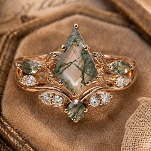 May include: A rose gold ring set featuring a large, kite-shaped moss agate gemstone. The ring is accented with smaller green gemstones and clear, round diamonds. The design includes leaf and floral details, creating an elegant and natural aesthetic.
