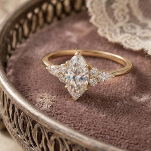 May include: A gold engagement ring with a marquise-cut center stone and smaller accent stones. The ring is set on a velvet surface inside a decorative silver tray, with a vintage paper and lace in the background.