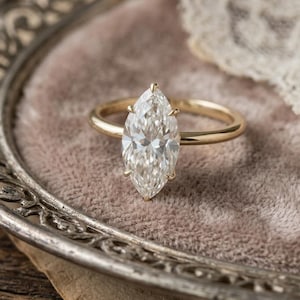 May include: A marquise-cut diamond engagement ring with a gold band. The diamond is set in a four-prong setting. The ring is displayed on a pink velvet surface, with a decorative silver tray and lace in the background.