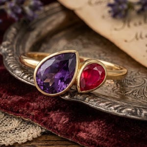 May include: A gold ring featuring two pear-cut gemstones set side-by-side. The larger stone is a deep purple amethyst, and the smaller stone is a vibrant red ruby. The band is polished gold, and both stones are secured in smooth gold bezels.