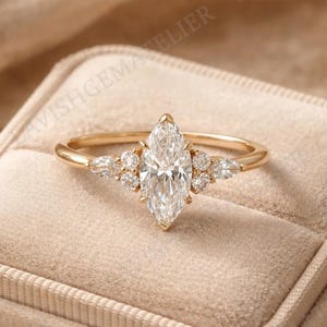 May include: A marquise-cut diamond engagement ring with a gold band. The ring features a large central diamond and smaller accent diamonds on either side. The ring is displayed in a beige velvet ring box.