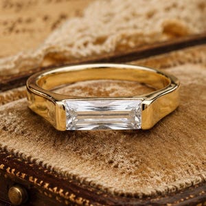 May include: A gold ring featuring a single horizontal baguette-cut clear gemstone set in a smooth metal band. The ring is positioned on a velvet-lined surface, showcasing the clarity of the stone and the polished finish of the gold metal band.
