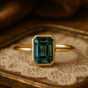 Może przedstawiać: Złoty pierścionek z prostokątnym, turkusowym kamieniem szlachetnym. Kamień jest osadzony w złotej oprawie, a pierścionek jest prezentowany na koronkowej powierzchni. Obrączka pierścionka jest prosta, polerowana.