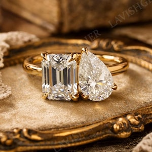 May include: A gold engagement ring featuring two large diamonds: an emerald-cut and a pear-shaped stone. The ring is set on a velvet surface within a decorative gold-toned frame, with lace in the background. The diamonds sparkle brightly.