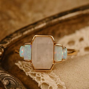 Może przedstawiać: Złoty pierścionek z dużym, prostokątnym, jasno zabarwionym kamieniem szlachetnym, otoczonym dwoma mniejszymi, prostokątnymi, opalizującymi kamieniami. Pierścionek leży na kremowej koronkowej serwetce.