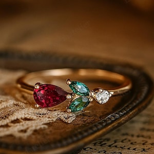 May include: A gold ring featuring a cluster of gemstones. The ring has a pear-shaped ruby, two emerald-cut emeralds, and a small diamond. The ring is set on a thin gold band, and the gemstones are arranged in a floral design.