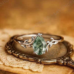 Puede incluir: Un anillo de plata con una piedra verde en forma de pera. El anillo presenta un delicado diseño de vid con hojas en la banda. El anillo se exhibe en un espejo de estilo vintage.