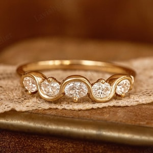 Puede incluir: Un anillo de oro con cinco diamantes de talla ovalada dispuestos en un patrón ondulado. El anillo se exhibe sobre un libro antiguo con una superposición de encaje, creando una estética cálida y elegante. El diseño del anillo es simple pero sofisticado.