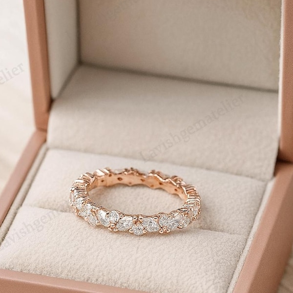 May include: A rose gold eternity ring with clear gemstones, presented in a pink jewellery box. The ring features a continuous row of sparkling stones, creating a full circle design. The box is open, revealing a soft, cream-coloured interior.