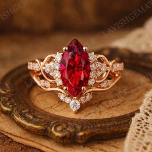 Op de afbeelding: Roségouden ring met een grote, marquise-geslepen, dieprode edelsteen als middelpunt. De ring is versierd met kleinere, ronde, heldere edelstenen en ingewikkelde filigraan details. De zetting is sierlijk en elegant, met een vintage uitstraling.