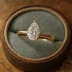 May include: A gold ring with a pear-shaped diamond set in a four-prong setting. The ring is displayed in a vintage-style, round, blue-green velvet ring box. The ring's band is thin and polished.