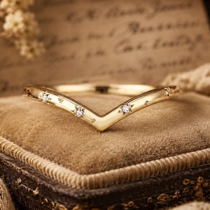 May include: A gold-colored chevron ring with embedded small diamonds. The ring is set on a brown velvet surface inside a vintage jewelry box. The ring's design includes star-shaped accents, adding a celestial touch to the jewelry.